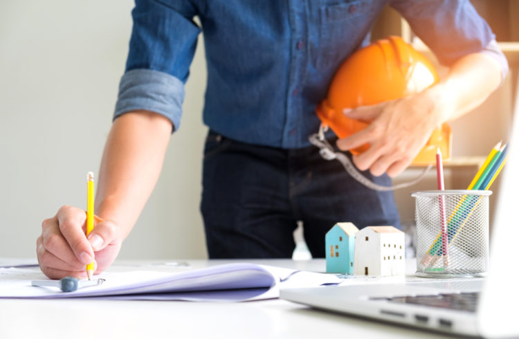 Architects Are Using Drawing Pencils,On The Other Hand Holds A Safety Hat,On The Writing Desk There Are Model Houses, Laptop And House Plans.