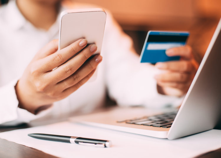 Close-up of female hand holding smartphone. Young businesswoman sitting at laptop in office holding credit card and entering data on her mobile phone