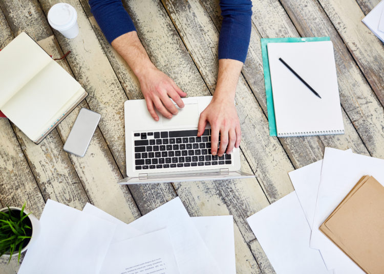 Top view of unrecognizable freelancer working on wooden floor, using laptop computer with documents and business supplies laid out around