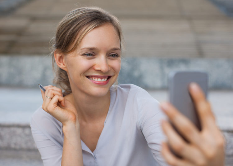 Portrait of cheerful young Caucasian businesswoman taking selfie or making video call with smartphone outdoors. Casual business and communication concept