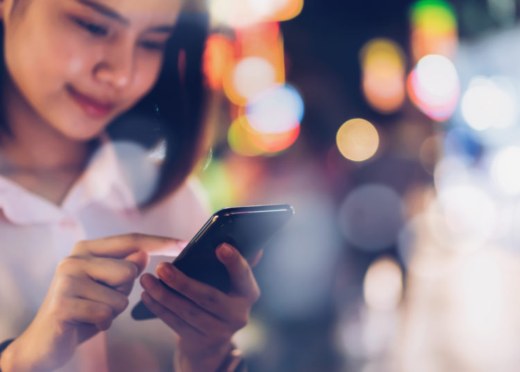 Closeup of woman holding a smartphone at night on city shopping street, and people walk. Technology for communication concept.Closeup of woman holding a smartphone at night on city shopping street, and people walk. Technology for communication concept.