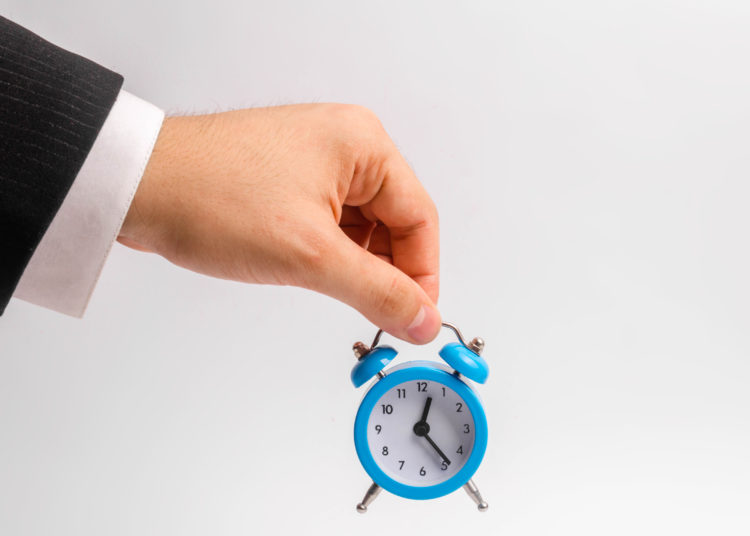 A businessman's hand holds a blue alarm clock on a white background. The concept of the flow of time, time to action. Hourly pay for work. Late for work and stay for part-time work. Time is money.