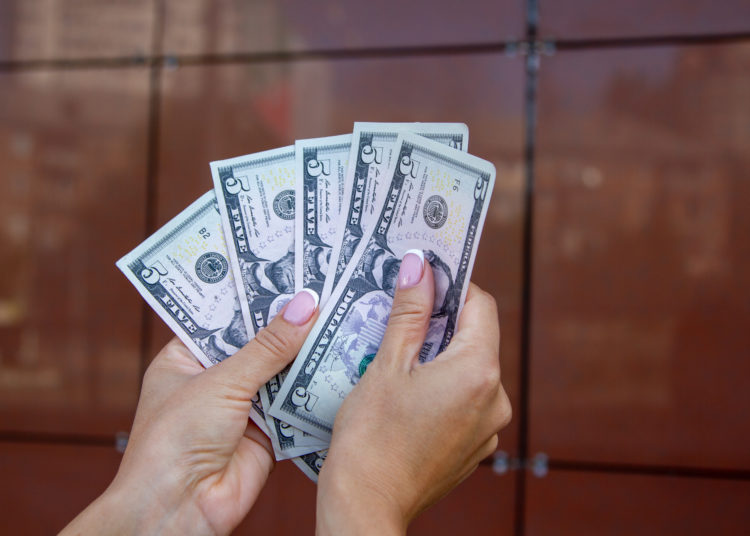 The woman counts the dollar bills, fanning them in her palms against the background of a brown shiny wall. Counting money. Crisp dollar bills.