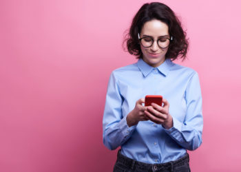 Portrait of a pretty smiling girl in summer clothes using mobile phone while standing isolated over pink background.