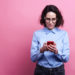 Portrait of a pretty smiling girl in summer clothes using mobile phone while standing isolated over pink background.