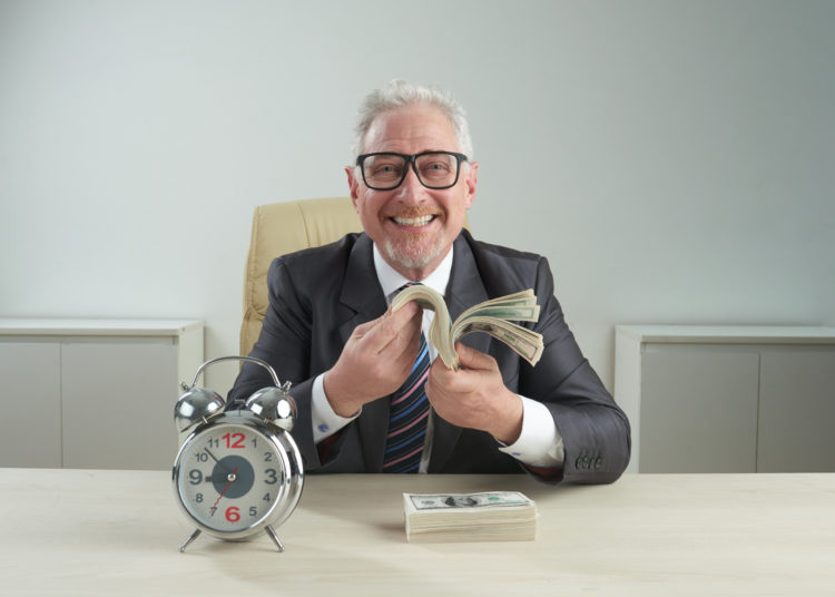 Cheerful senior entrepreneur wearing classical suit and eyeglasses looking at camera with toothy smile while holding bundle of banknotes in hands, alarm clock standing on office desk