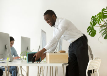 Happy african new employee unpacking box with belongings at workplace on first working day concept, excited black hired worker newcomer smiling holding framed picture settling in office on desk
